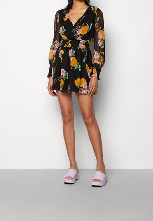 Kvinde iført en sort blomsterprintet minikjole med lange gennemsigtige ærmer og lilla platformsandaler stående mod en ensfarvet hvid baggrund.