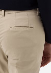 Beige broek met een achterzak met een platte sluiting, een gladde textuur en een subtiele geborduurde merklogo dicht bij de zak.