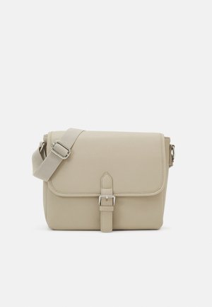 Bolso bandolera beige con correa de tela ajustable y solapa frontal asegurada con una correa y hebilla plateada, sobre un fondo blanco.