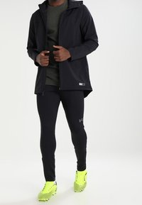 Černá voděodolná bunda s kapucí, doplněná těsnými černými legínami. Jasně žluté kopačky s texturovaným vzorem pro lepší přilnavost.