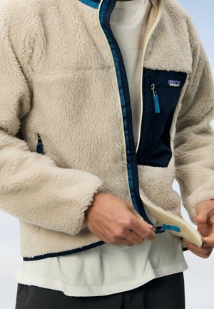 Person trägt eine beige Fleecejacke mit marineblauer Brusttasche und Besatz, gezeigt beim Schließen des vorderen Reißverschlusses über einem weißen Hemd.