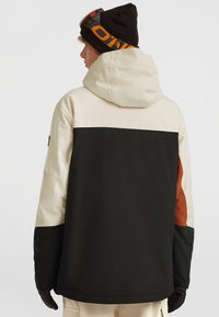 Waterdichte jas met een beige bovenstuk, zwart onderstuk en oranje accenten. Beschikt over een capuchon en een gestructureerde stof voor isolatie.