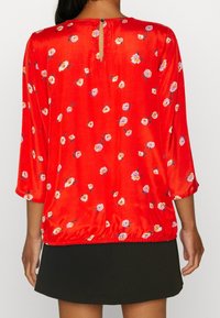 Blusa de satén roja con mangas de tres cuartos, con un estampado floral en rosa y blanco. Detalle de abertura en la parte posterior con un botón. Dobladillo fruncido.