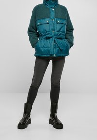 Tyrkysová zateplená bunda s texturovanou fleecovou částí v horní části, dvěma předními kapsami, nastavitelným pasem a patentními zápichy. Nošena s tmavými skinny džíny a černými botami.