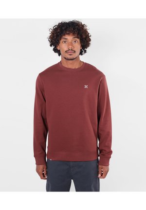 Homme aux cheveux bouclés portant un sweat-shirt uni bordeaux à manches longues et un pantalon foncé, debout devant un fond clair.