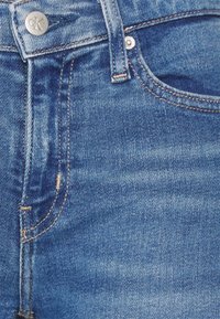 Primer plano de unos vaqueros de mezclilla azul que muestra la cinturilla, el botón y las costuras del bolsillo, resaltando la textura y el color de la tela.