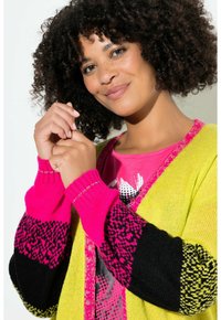 Strickjacke mit leuchtend gelbem Körper, Ärmeln mit pink-schwarzem Muster und gerippten Bündchen. Getragen über einem pinken Grafikshirt.