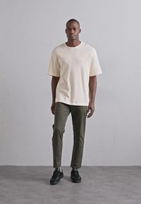 Beige strukturiertes Kurzarmhemd, olivgrüne Hose, schwarze Sneakers. Das Model steht vor einem schlichten hellen Hintergrund.