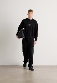 Zwarte sweatshirt met een vierkant patch, zwarte joggingbroek en zwarte schoenen. Het model draagt een grote, ronde zwarte tas. Simpel, monochroom ontwerp.