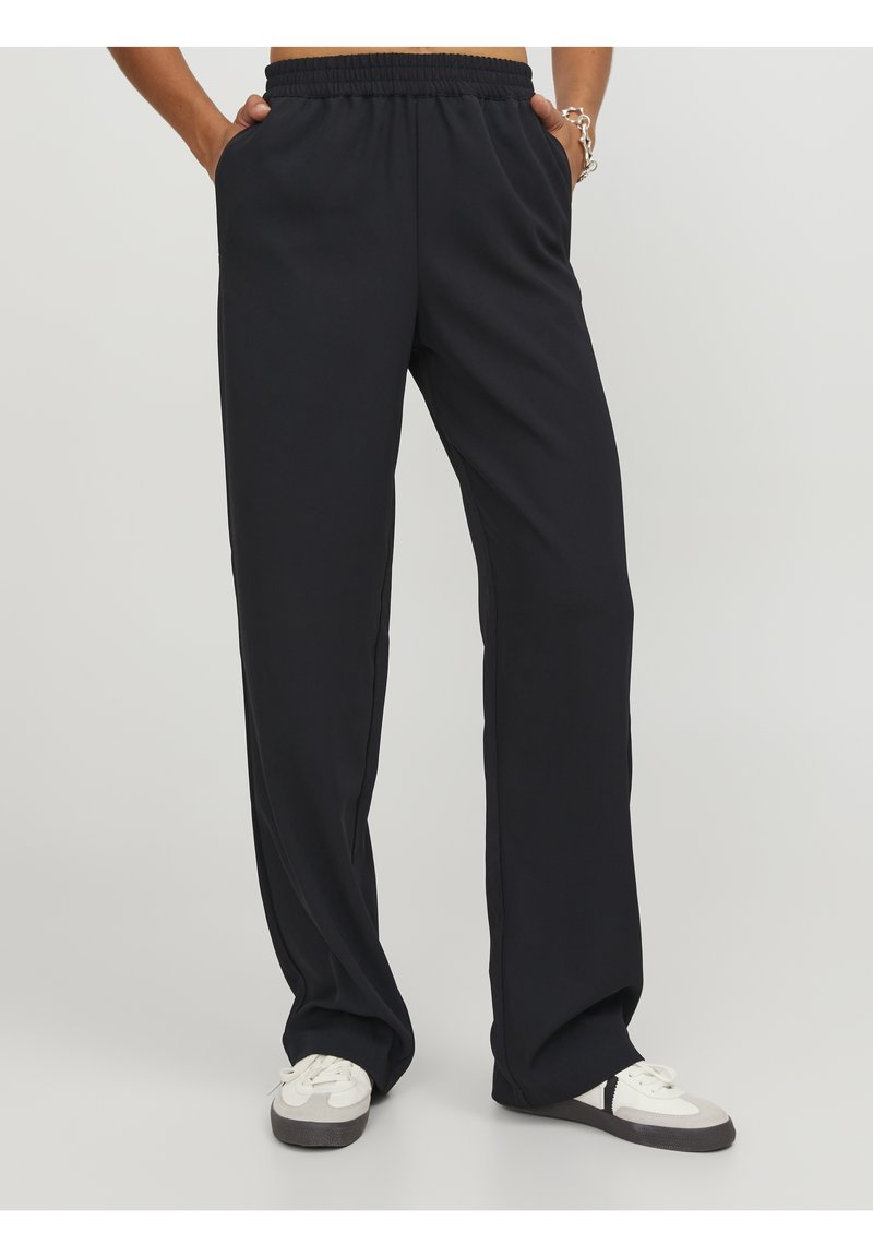 Personne portant un pantalon noir ample avec une ceinture élastique et des baskets blanches, debout les mains dans les poches sur un fond uni.