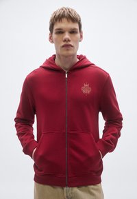 Červená mikina na zips s predným vreckom, na hrudi je jemné logo, vyrobená z mäkkej, štruktúrovanej látky s kapucňou so šnúrkou.