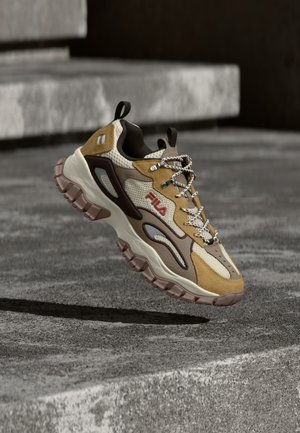 Χοντρό sneaker FILA με μπεζ, καφέ και ταμπά πάνελ, παχιά ανθεκτική σόλα, κορδόνια με μοτίβο και θηλιές, αιωρούμενο πάνω από υφή τσιμεντένιων σκαλοπατιών.