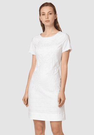 Mujer con un vestido blanco de manga corta con patrones florales texturizados y bordes festoneados, de pie frente a un fondo claro y liso.