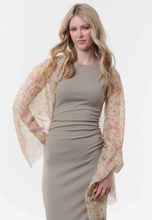 Donna bionda con capelli mossi indossa un vestito beige aderente e un foulard floreale trasparente drappeggiato sulle braccia, su sfondo bianco.