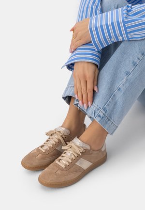 Osoba nosząca beżowe zamszowe trampki z białymi akcentami, jasnoniebieskie dżinsy i niebiesko-białą koszulę w paski, z rękami opartymi na kolanie.