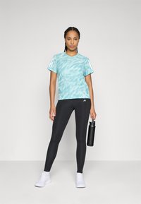 Donna in piedi con una maglietta turchese a fantasia, leggings neri, sneakers bianche, mentre tiene una bottiglia d'acqua nera contro uno sfondo semplice.