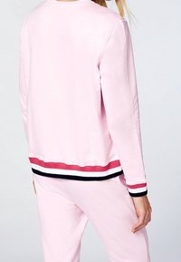 Femme portant un sweat-shirt rose clair et un pantalon assorti avec des bordures rayées rouge, blanc et noir aux poignets et à l'ourlet, vue de dos.