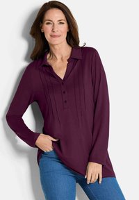 Blusa a maniche lunghe, con bottoni, di un profondo viola con pieghe frontali e colletto. Indossata con jeans blu. Tessuto morbido, vestibilità rilassata.