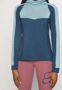 Långärmad tröja i mörkblått och ljusblått med hög krage. Har kontrasterande färgblock och en logotyp på bröstet. Slät tygfinish.