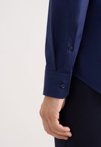 Camicia a maniche lunghe blu navy con una texture liscia, polsini arrotondati con due bottoni e taglio sartoriale, mostrata dal gomito al polso.