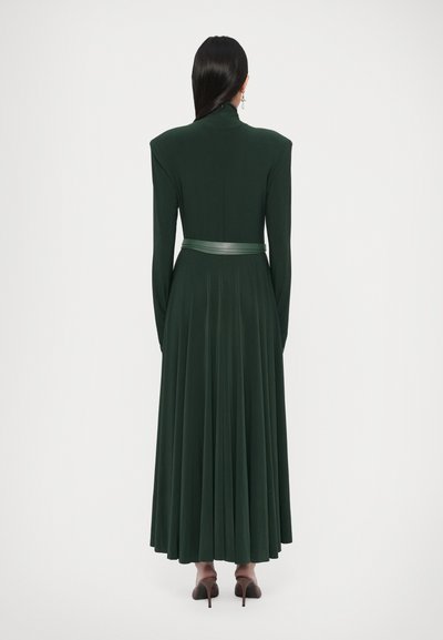 Gara, tumši zaļa kleita ar piegulošu augšdaļu un plūstošu kroku svārku. Ar polsterētiem pleciem un atbilstošu jostu viduklī.