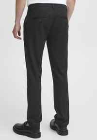 Schwarze Hose aus glattem Stoff mit taillierter Passform, zwei Gesäßtaschen mit Knöpfen und kombiniert mit schwarzen Slippern.