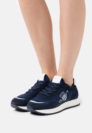 Para granatowych sneakersów GANT z białym logo i podeszwą noszonych na bosych stopach na białym tle.