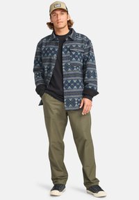 Button-up flannel shirt featuring blue and green geometric patterns, paired with olive pants and black sneakers with white soles.