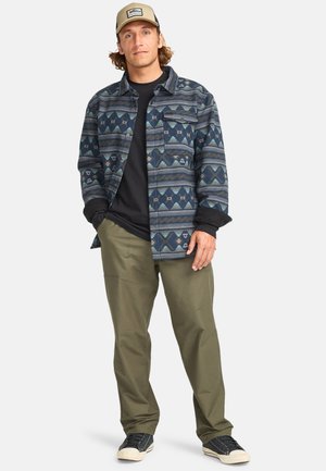 Button-up flannel shirt featuring blue and green geometric patterns, paired with olive pants and black sneakers with white soles.