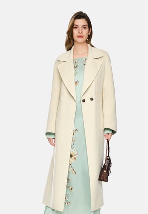 Woman wearing a long cream coat over a light blue floral dress, holding a small brown handbag with a thin strap.