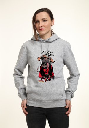 Femme portant un sweat à capuche gris avec un chien de dessin animé déguisé en Deadpool imprimé à l'avant, debout les mains détendues, fond neutre.