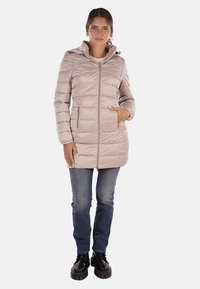 Cappotto imbottito beige con zip frontale, cappuccio e tasche laterali. Presenta una texture liscia e dettagli di cucitura sottili. Abbinato a jeans e stivali.