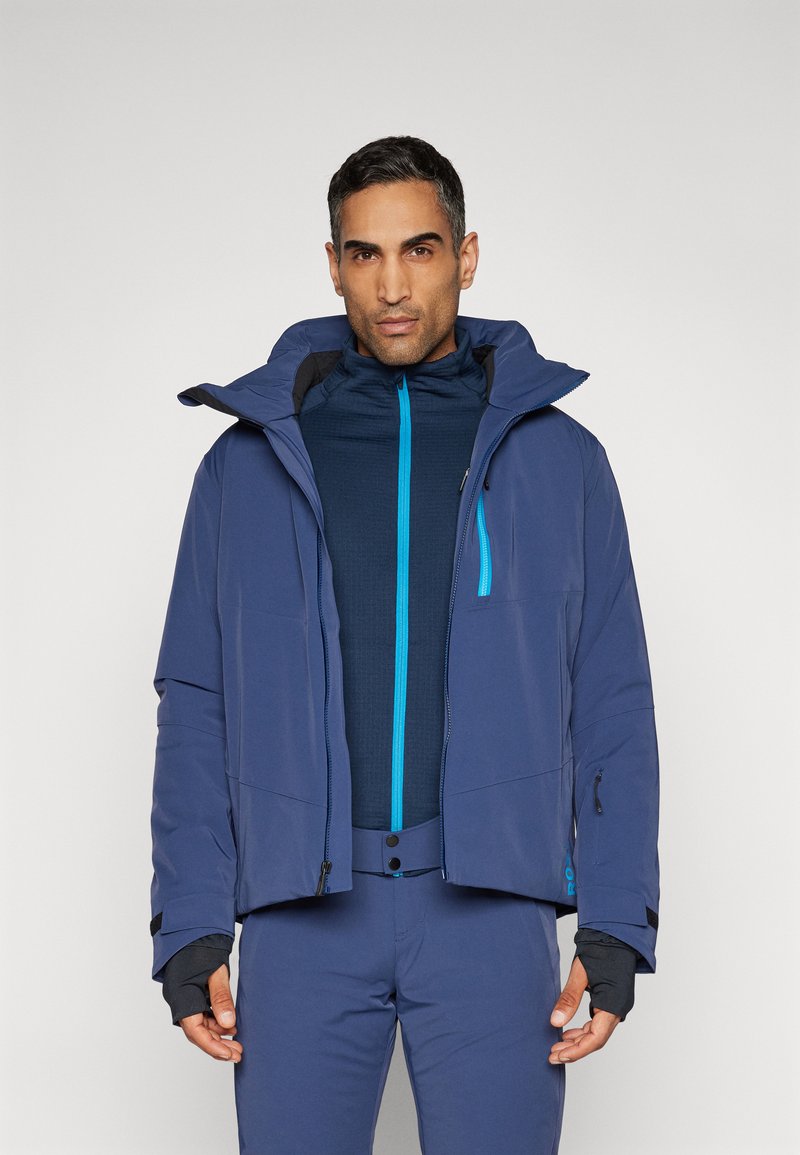 Veste de ski bleu marine avec col haut, capuche ajustable et fermeture éclair en teal contrastant. Présente un tissu texturé et des poches zippées.