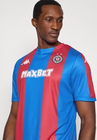 Μπλε και κόκκινη ριγέ φανέλα ποδοσφαίρου κατασκευασμένη από αεριζόμενο ύφασμα, με λαιμόκοψη κανονική και το λογότυπο "MAXBET" να προβάλλεται έντονα στο μπροστινό μέρος.