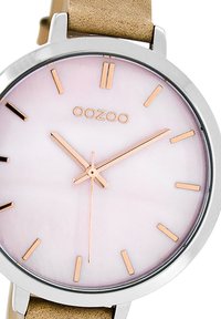Montre-bracelet en or rose avec un boîtier rond en argent, un cadran rose et un bracelet en cuir tan. Présente des marqueurs horaires et des aiguilles minimalistes.