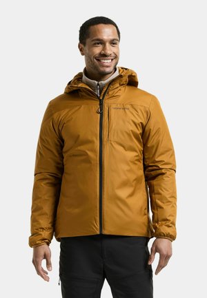 Smiling man wearing a mustard yellow hooded jacket with front zipper and black pants, standing against a plain background.