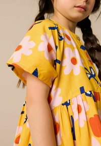 Keltainen mekko, jossa on suuria vaaleanpunaisia ja oransseja kukkia, lyhyet hihat, nappikiinnitys edessä ja rypytetty vyötärö, sinisillä yksityiskohdilla.