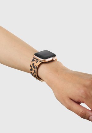 Poignet portant une montre connectée dorée avec écran noir et bracelet à motif léopard sur un fond clair uni.