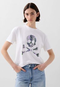 Hvid bomulds t-shirt med et grafisk design af dødningehoved og korslagte knogler i lilla og grøn, korte ærmer og rund halsudskæring.