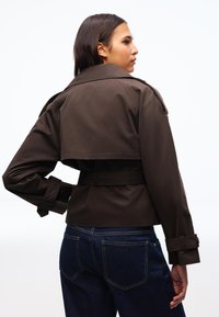 Giovane donna con capelli lunghi che indossa una giacca corta marrone scuro con cintura e jeans blu, posando con una mano sul fianco.