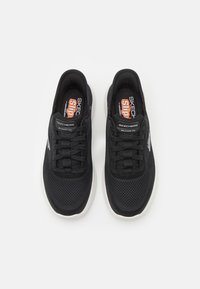 Zapatillas deportivas negras con parte superior de malla, diseño de cordones y suela blanca. Presenta acentos de marca en naranja y gris en la lengüeta y el costado.