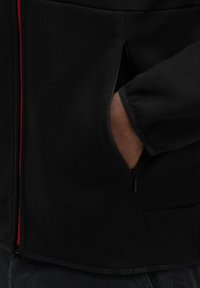 Giacca nera con zip e dettagli rossi, dotata di tasca laterale e tessuto testurizzato, taglio aderente e un logo discreto sull'orlo.