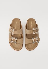 Tan mockasinsandaler med två breda remmar, var och en med en juvelprydd fyrkantig spänne, på en platt brun sula.