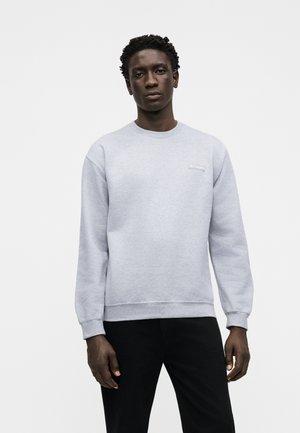 Jeune homme portant un sweat-shirt gris clair à manches longues et un pantalon noir, debout devant un fond blanc uni.