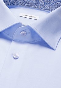 Chemise bleue claire en tissu lisse et texturé. Comprend un col classique, deux boutons blancs et un col intérieur bleu à motifs.