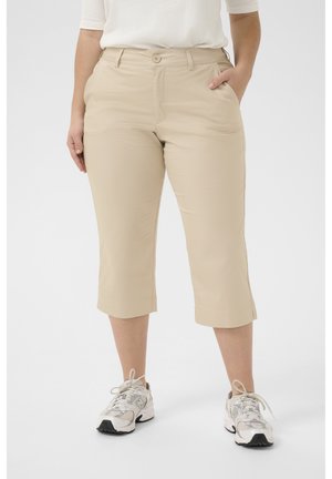 Femme portant un pantalon beige court et des baskets blanches, debout avec une main dans la poche devant un fond uni.