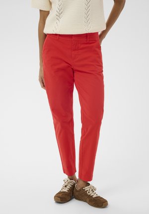 Persoană care poartă pantaloni conici de culoare roșu aprins, asortați cu adidași maro cu șireturi și un top fără mâneci din tricot crem.