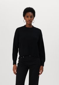 Maglione nero lavorato a maglia con una texture a coste, scollo rotondo e orlo leggermente accorciato. Indossato con pantaloni neri, con maniche lunghe e una vestibilità rilassata.