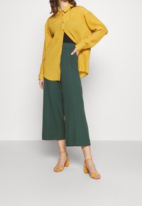 Nainen, joka pitää yllään löysää keltaista nappipaitaa, tummanvihreitä leveälahkeisia housuja ja keltaisia nilkkaremmillä varustettuja korkokenkiä, seisoo yksinkertaista taustaa vasten.