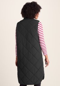 Woman facing away wearing a long quilted black vest over a pink and white striped long-sleeve shirt against a plain light background.